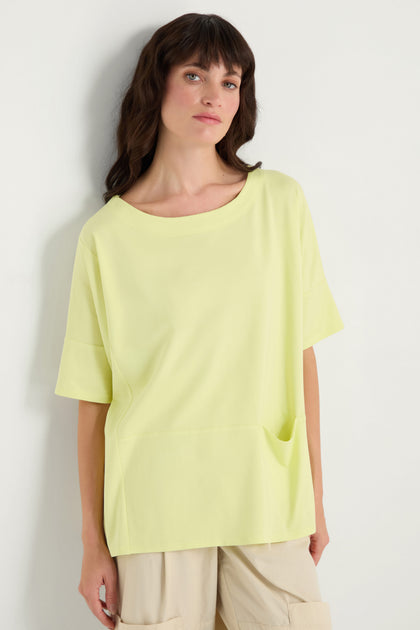 A person wears the Jersey Pocket Top, a relaxed fit light yellow viscose jersey with a small front pocket, paired with beige pants, standing against a white background.