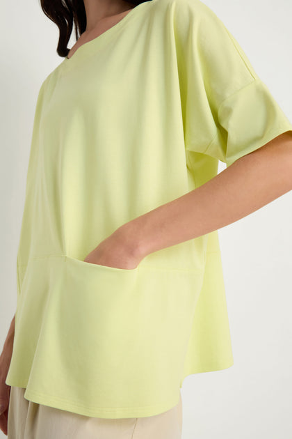A person wearing the Jersey Pocket Top, a loose, relaxed fit light yellow viscose jersey with one hand in its large front pocket.