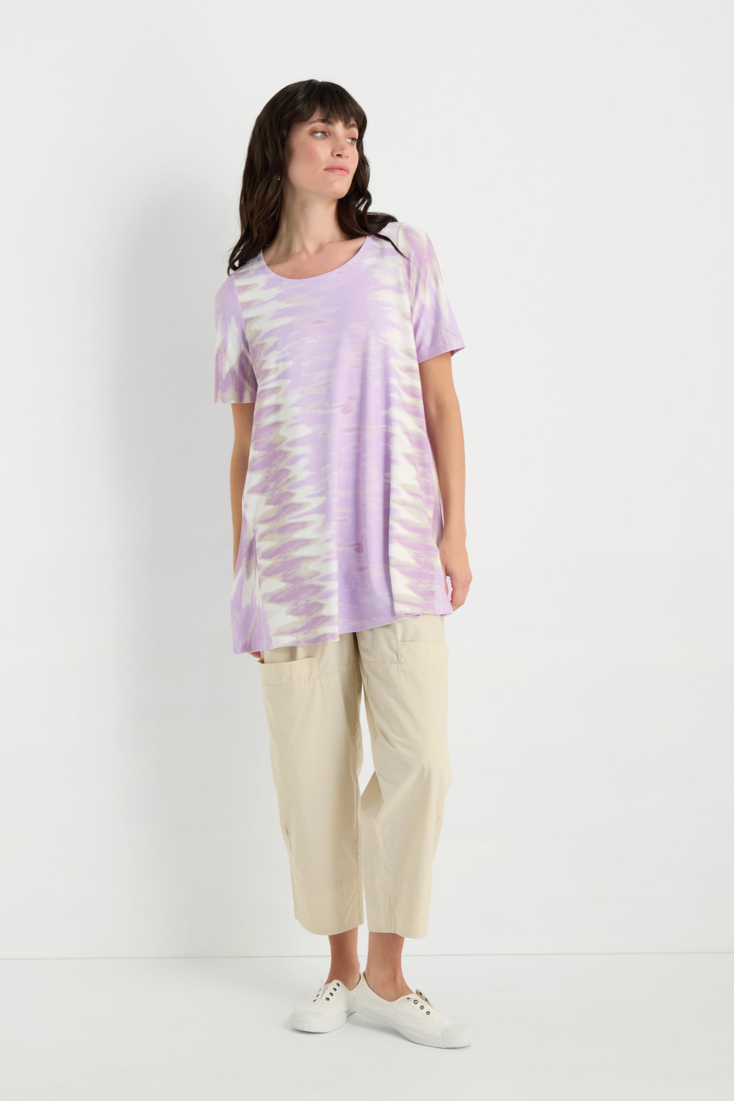 A woman stands against a white background wearing the Tie-Dye Jersey Tunic in loose lavender style, paired with beige cropped pants and white sneakers.