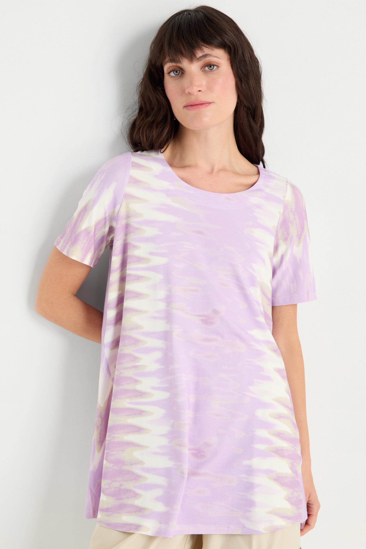 A woman wears the Tie-Dye Jersey Tunic, a relaxed fit top in lavender and cream with short sleeves and tie-dye details, standing against a plain white wall.