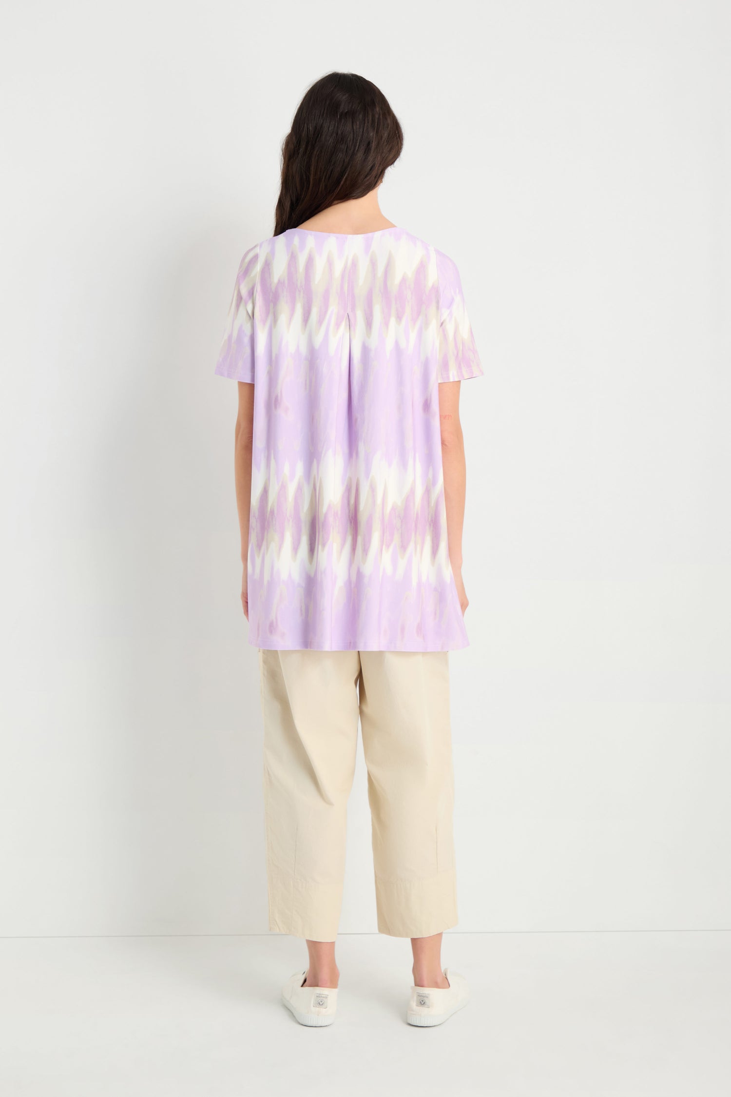 A person stands facing away, wearing the Tie-Dye Jersey Tunic in loose lavender and white, paired with beige wide-leg pants and white shoes against a plain white background.