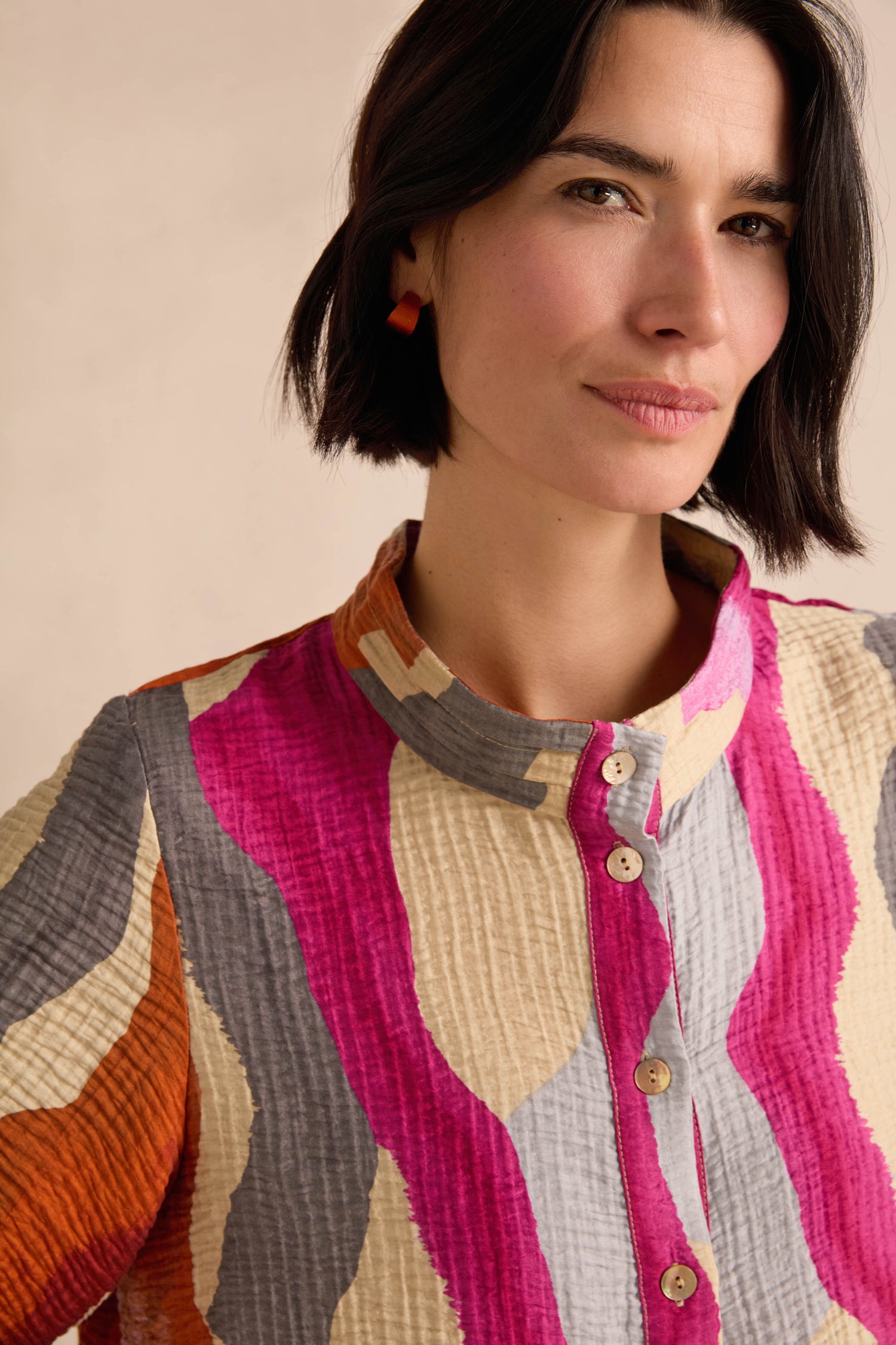 A woman with short dark hair stands against a neutral background, wearing the Wavy Painted Lines Shirt—a crinkled viscose top adorned with pink, orange, gray, and beige wavy patterns and finished with wooden buttons.