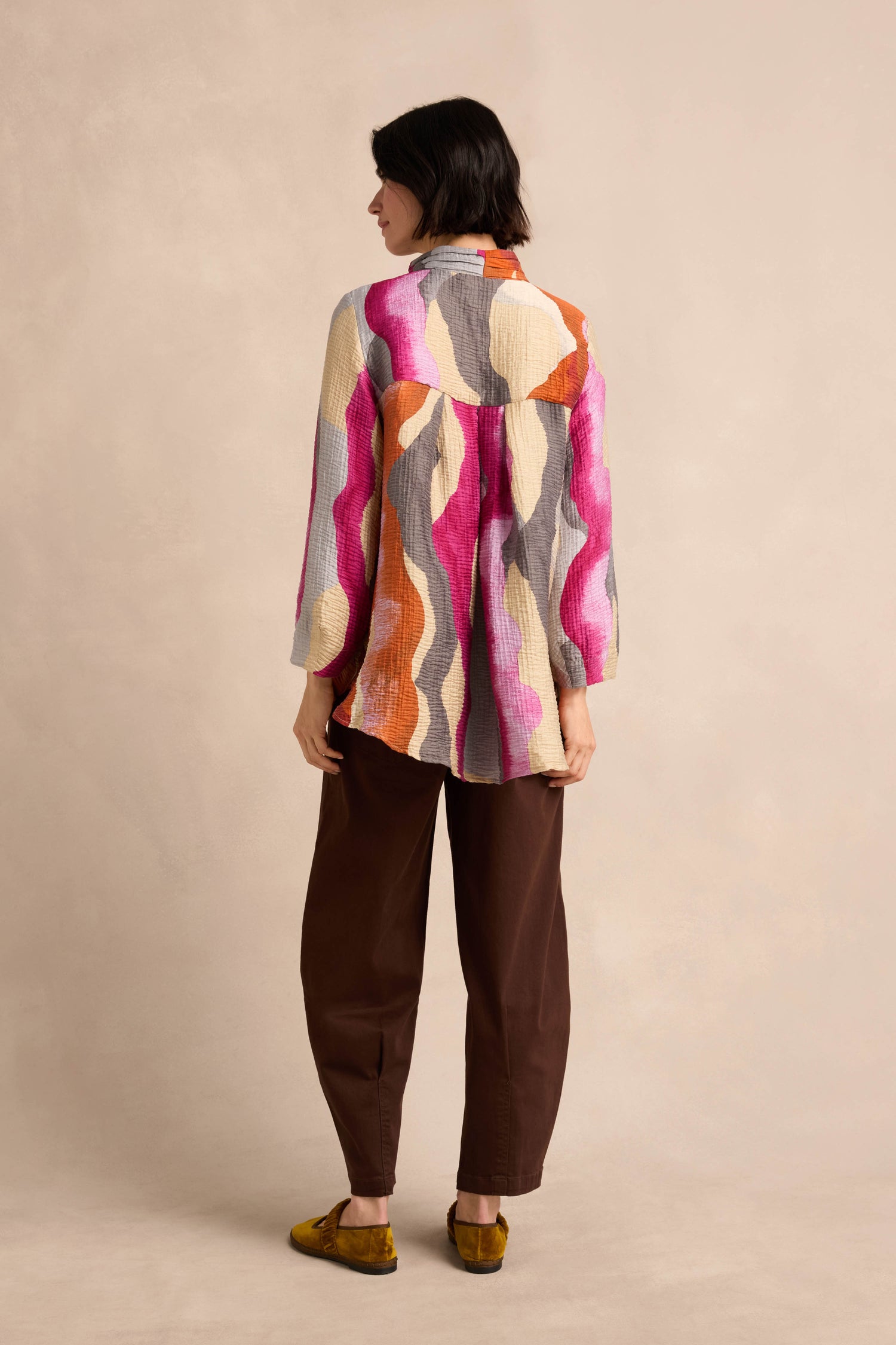 A person stands with their back to the camera, wearing the Wavy Painted Lines Shirt—a colorful crinkled viscose top featuring abstract patterns—paired with brown pants and mustard yellow shoes, set against a neutral background.