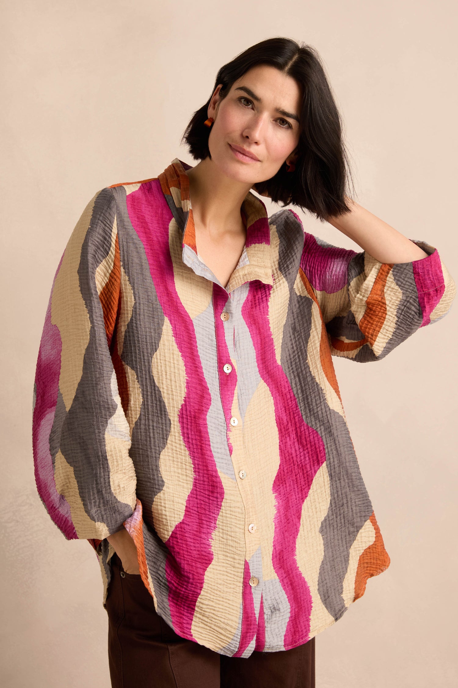 A woman wearing the Wavy Painted Lines Shirt—a loose, longline shirt featuring pink, beige, orange, and gray wavy stripes—stands against a plain light background.