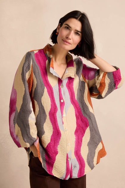 A woman wearing the Wavy Painted Lines Shirt—a loose, longline shirt featuring pink, beige, orange, and gray wavy stripes—stands against a plain light background.