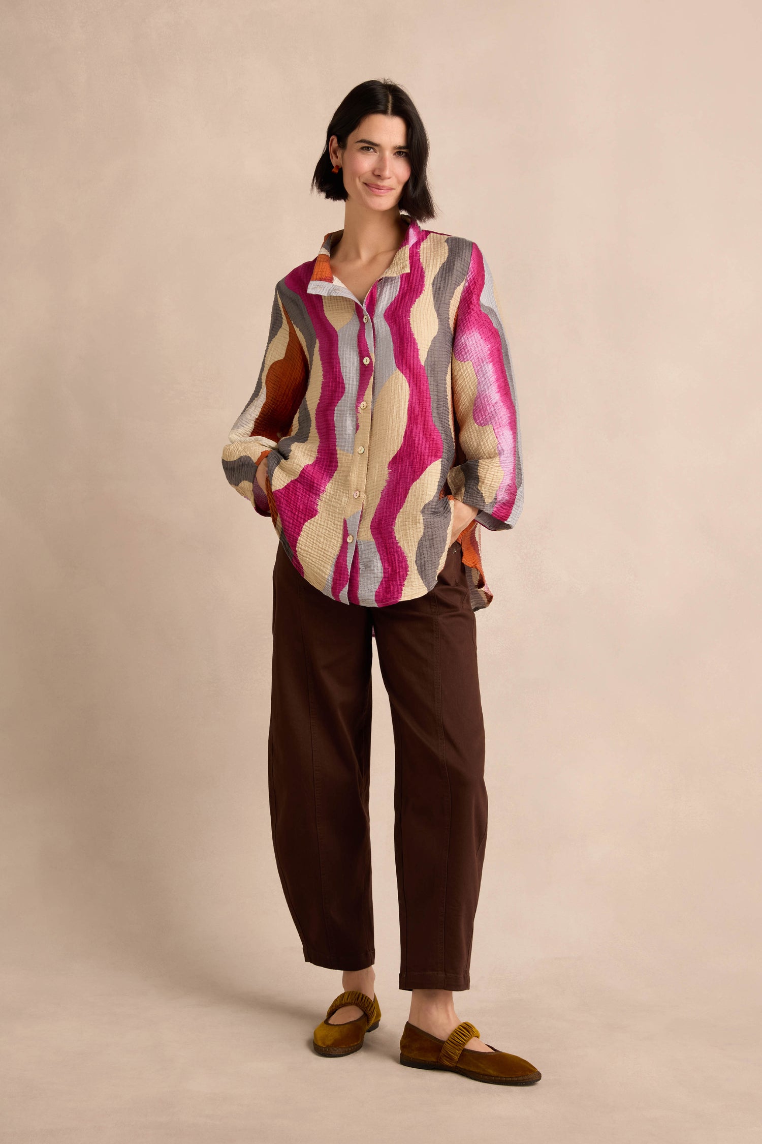 A woman stands against a beige background, hands in pockets and a slight smile on her face, wearing the Wavy Painted Lines Shirt styled as a button-up, paired with brown pants and yellow shoes.