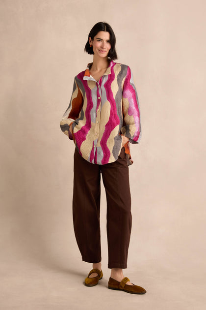 A woman stands against a beige background, hands in pockets and a slight smile on her face, wearing the Wavy Painted Lines Shirt styled as a button-up, paired with brown pants and yellow shoes.