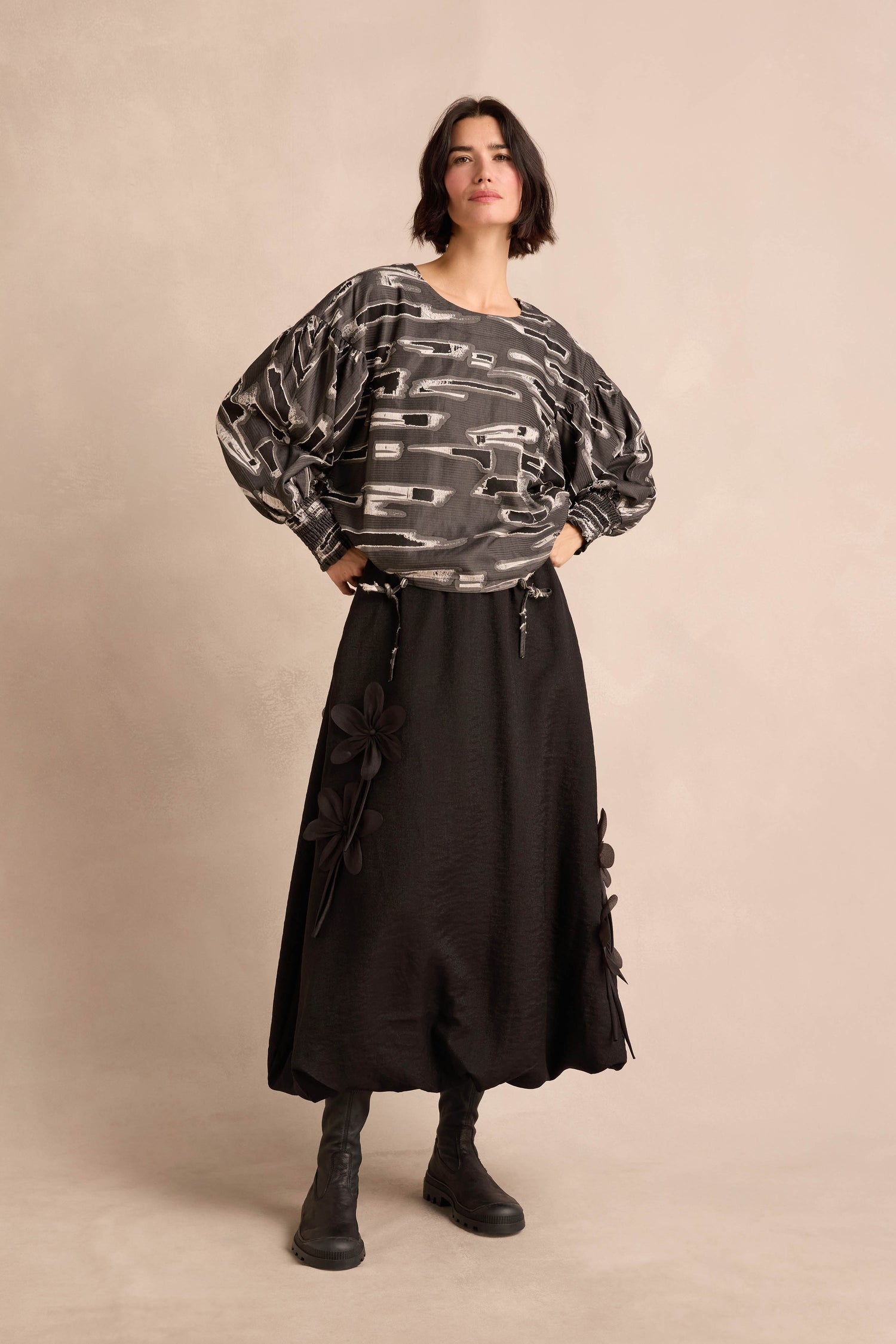 A woman stands against a neutral background wearing the Abstract Embroidery Top, styled with a black ankle-length skirt decorated with floral appliqués and black boots, hands on hips.