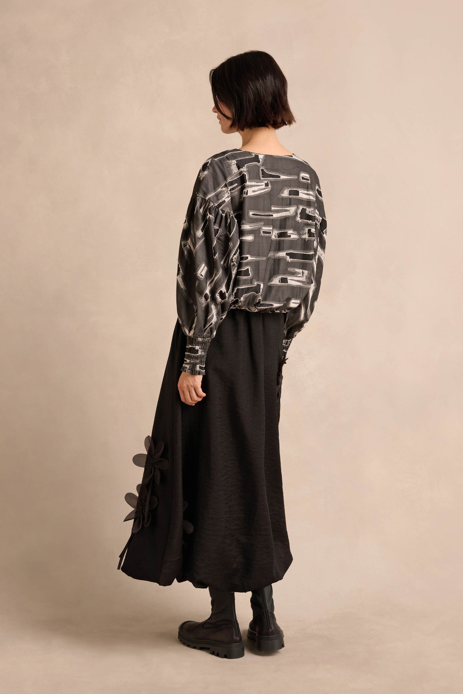 A person facing away, wearing the Abstract Embroidery Top—a patterned gray and black blouse with graphic embroidered blocks—paired with a long black skirt featuring floral details and black boots, standing against a neutral background.