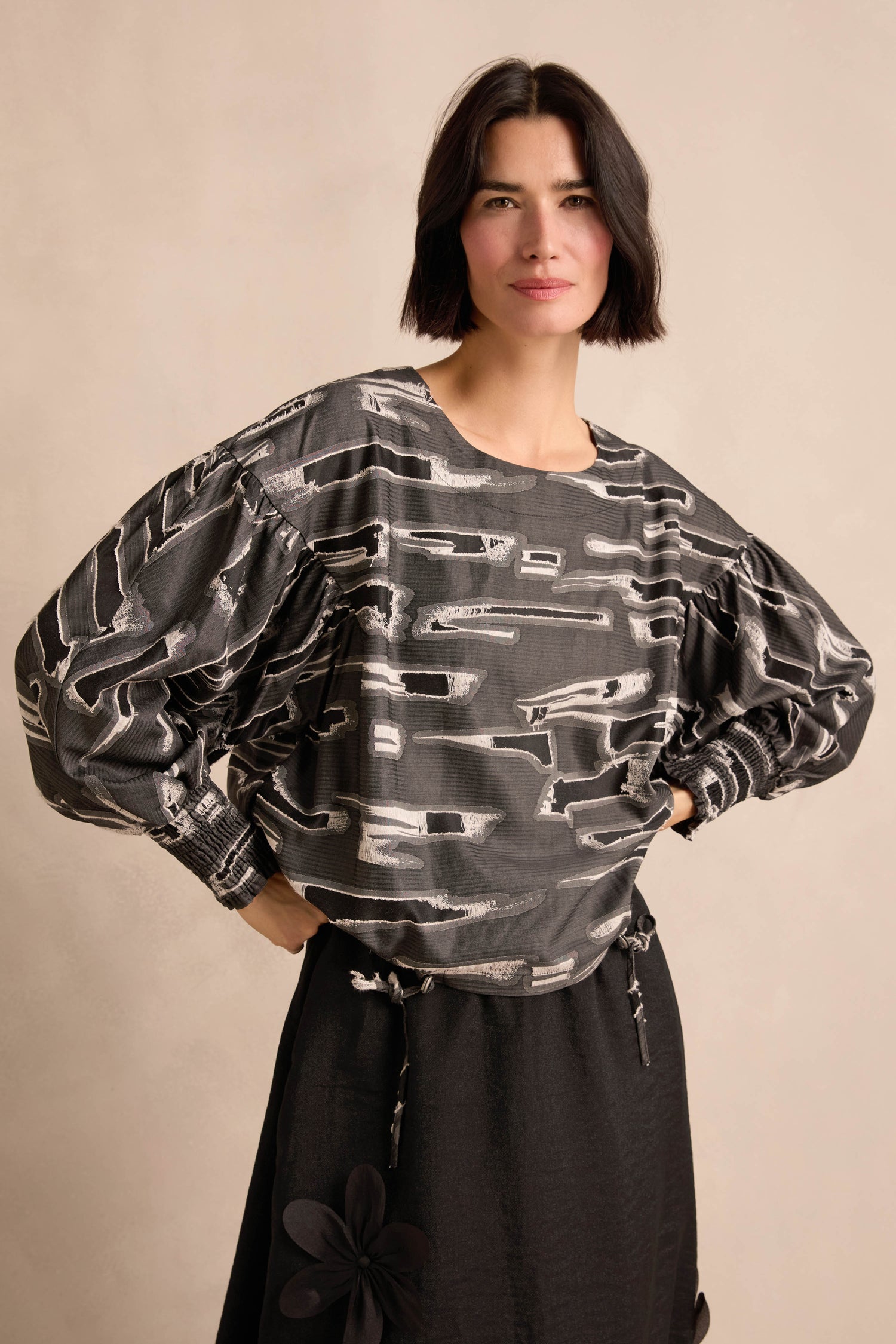 A woman with short dark hair stands with her hands on her hips against a neutral background, wearing the Abstract Embroidery Top in black and gray paired with a black skirt.