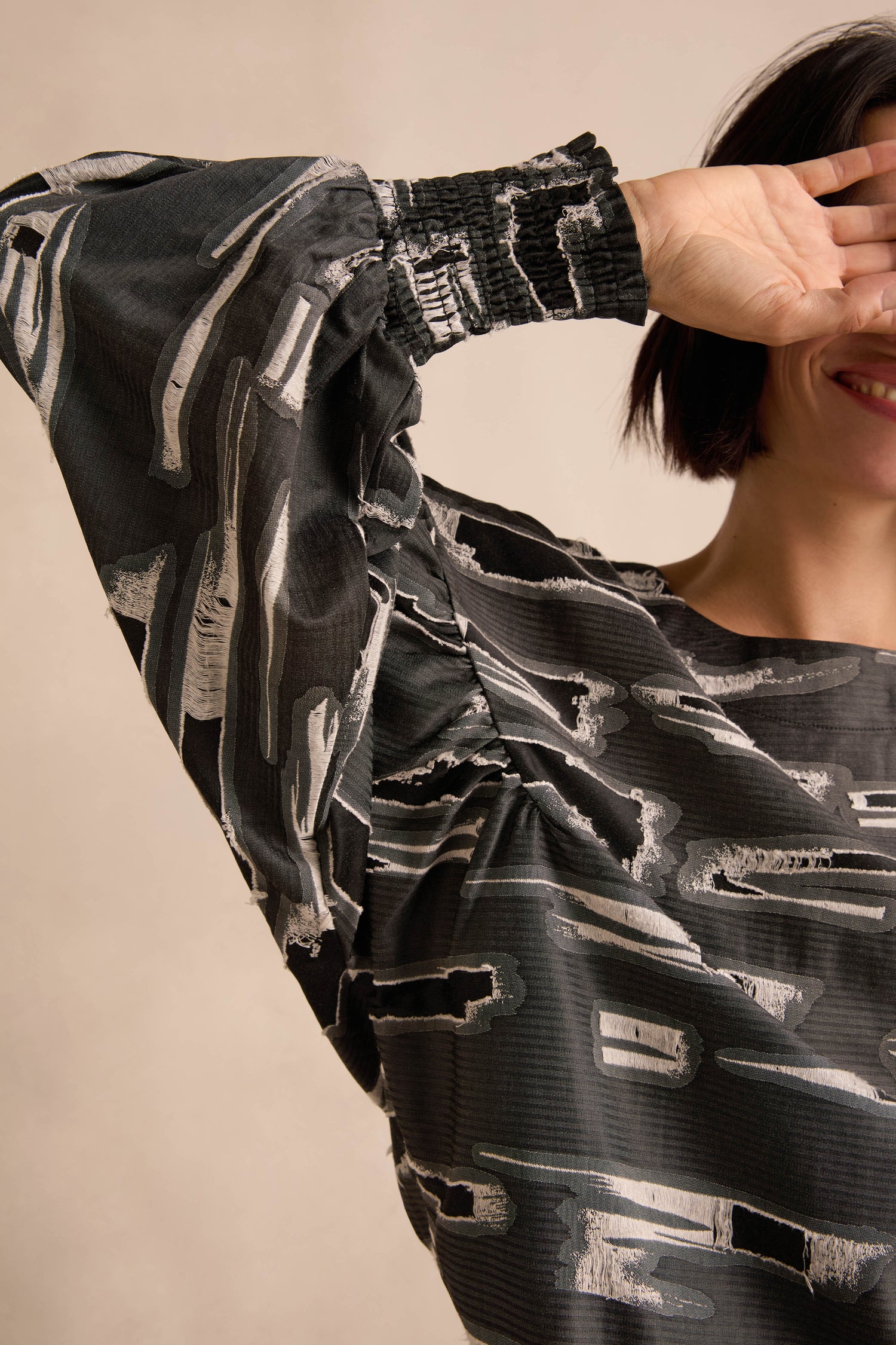 Wearing the Abstract Embroidery Top featuring a black and gray abstract pattern, a woman with short dark hair smiles as she raises her arm, partially covering her face with her hand.