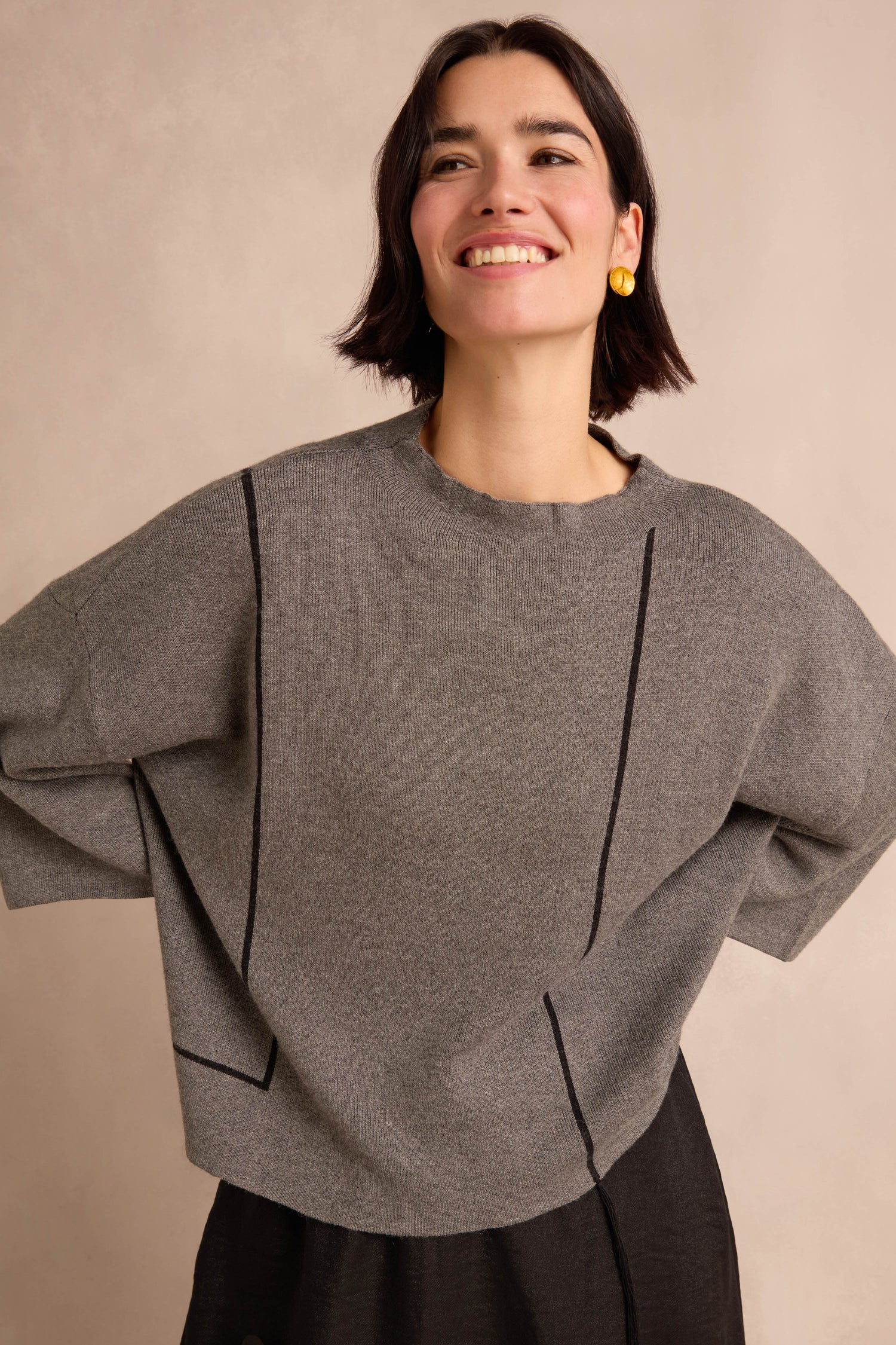 Woman smiling, wearing the Piping Detail Knit—a relaxed fit gray sweater with black lines—paired with a black skirt and yellow earrings, standing against a neutral background.