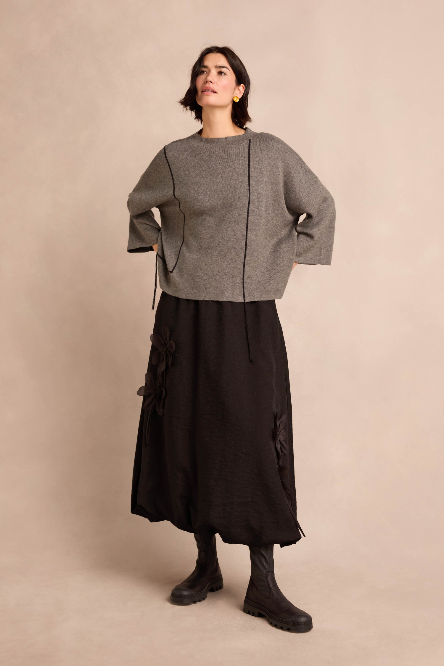 A woman stands against a beige background, wearing the Piping Detail Knit in grey, paired with a long black skirt featuring floral details and black boots.