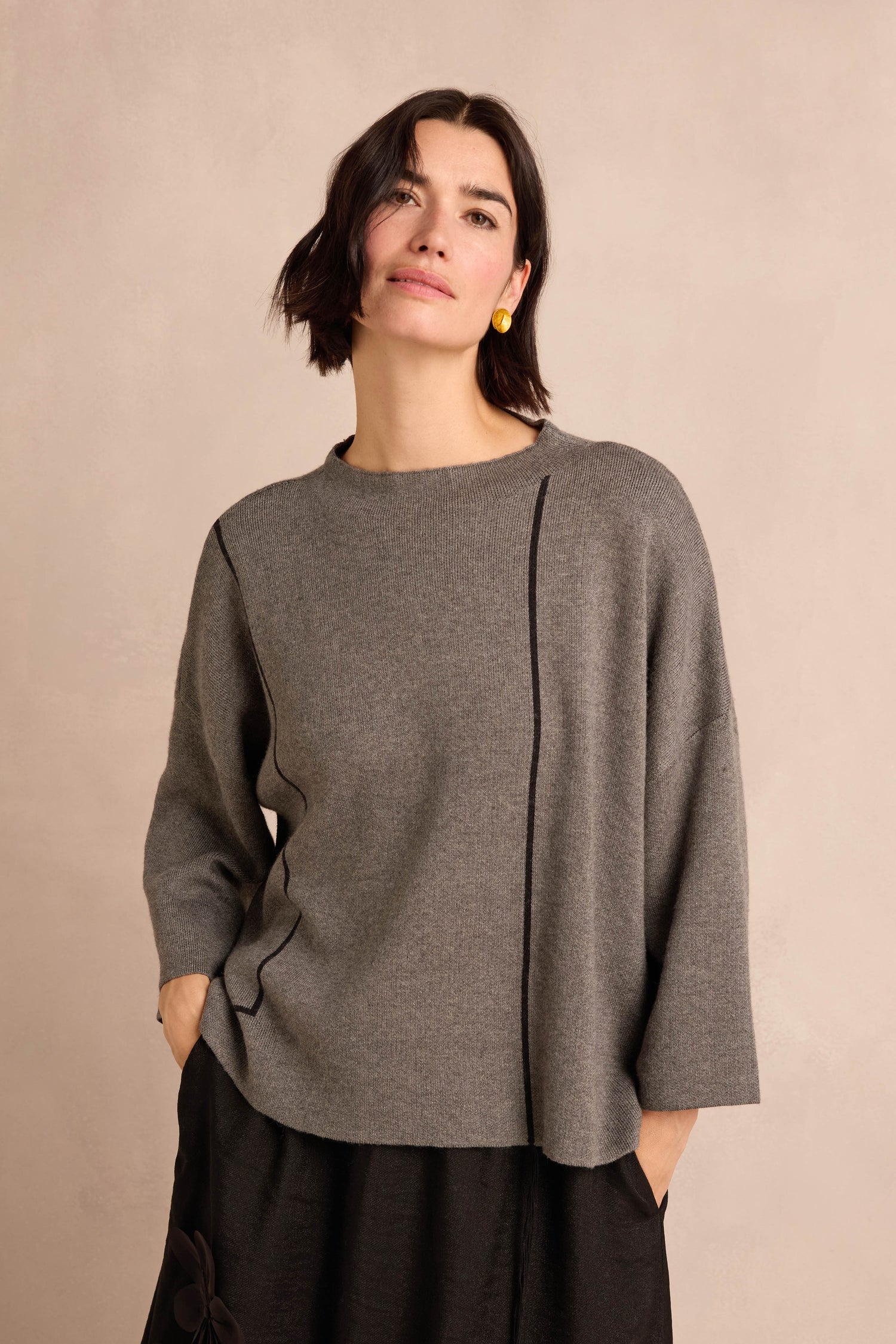 A woman wearing the Piping Detail Knit, a relaxed fit gray wool-enriched sweater, and a black skirt stands against a neutral background with one hand in her pocket.