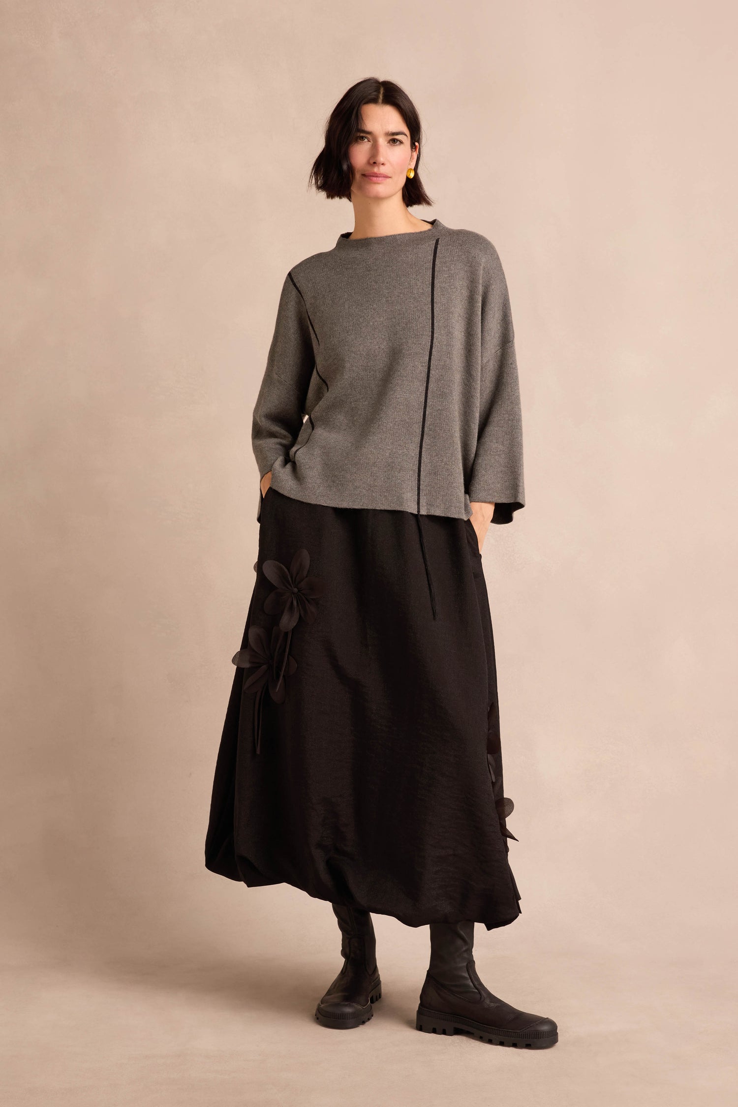 A woman wears the Piping Detail Knit in a loose, relaxed fit paired with a long black skirt featuring floral accents and black boots, standing against a plain beige background.
