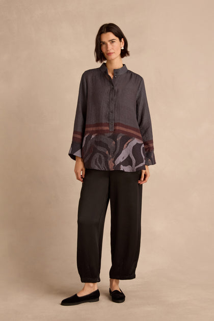 A woman stands against a beige background, wearing the Print Hem Pocket Shirt featuring an abstract border print, paired with black pants and black shoes.