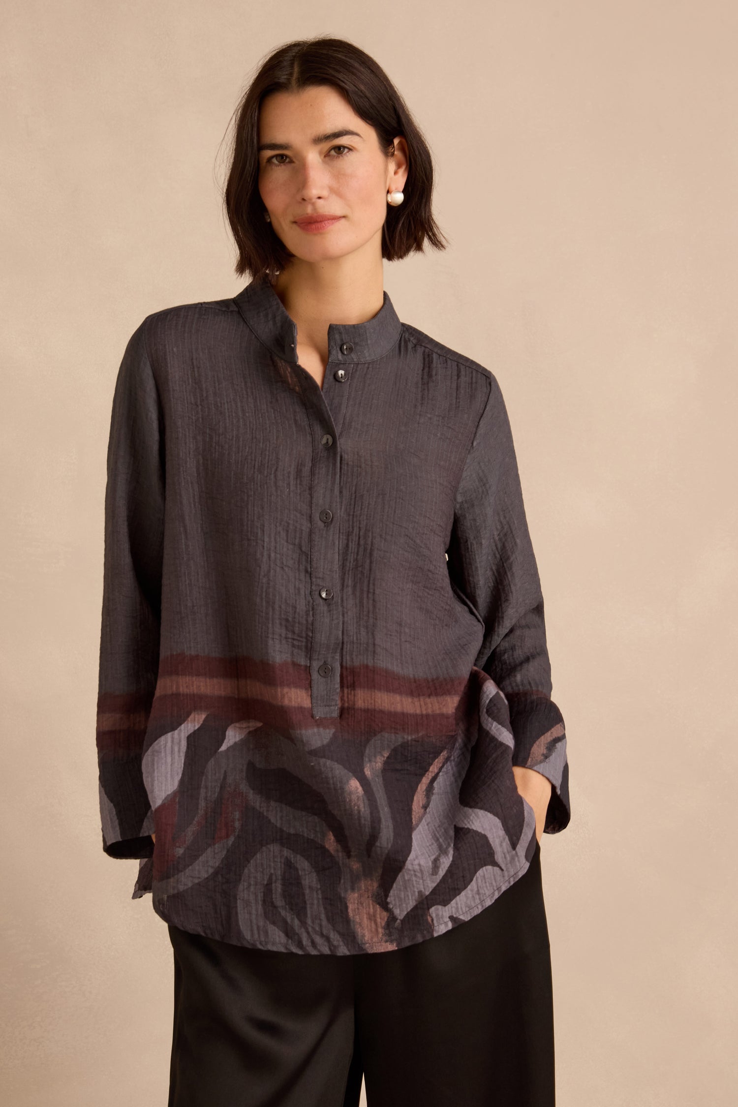 A woman with short dark hair wears the Print Hem Pocket Shirt—a relaxed fit shirt featuring an abstract border print—paired with black pants, standing against a plain beige background.
