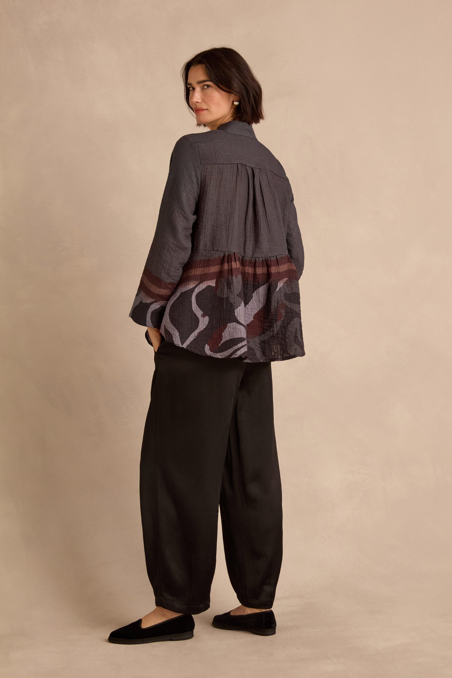 A person stands against a beige background, dressed in the Print Hem Pocket Shirt—a loose, relaxed fit shirt with a red and gray abstract border print on the back—paired with black pants and black shoes.