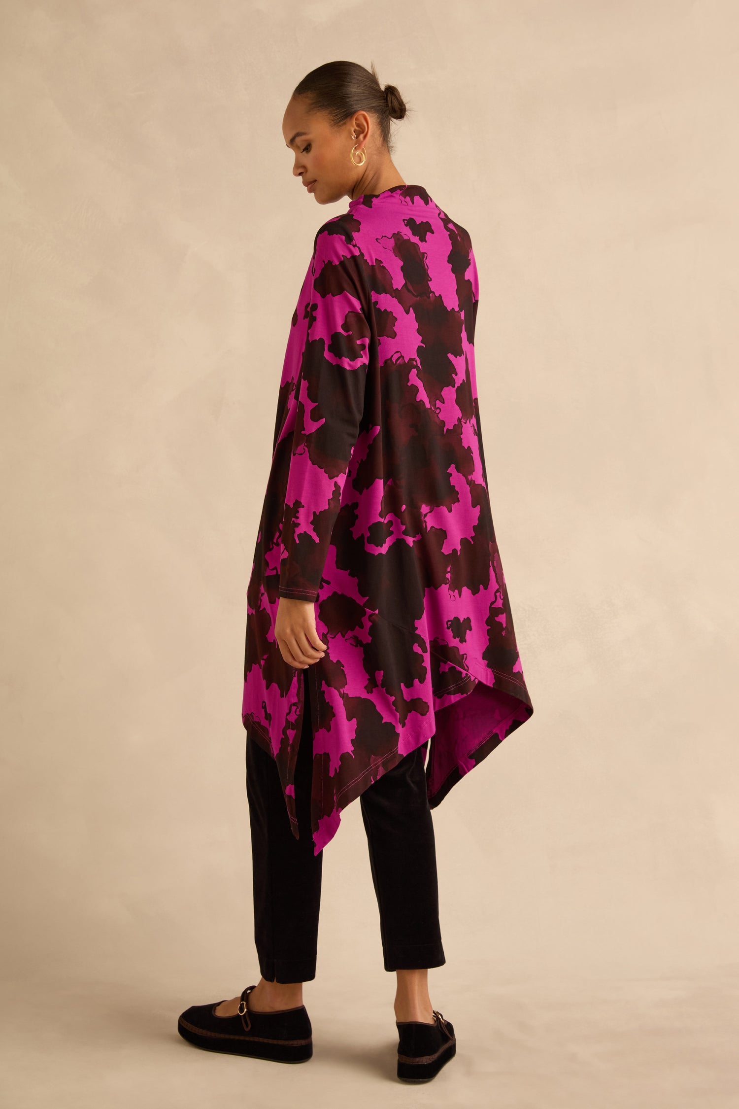 A woman wearing an Autumn Leaves tunic with a long, asymmetrical pink and brown abstract botanical print, paired with black trousers and dark buckled shoes.