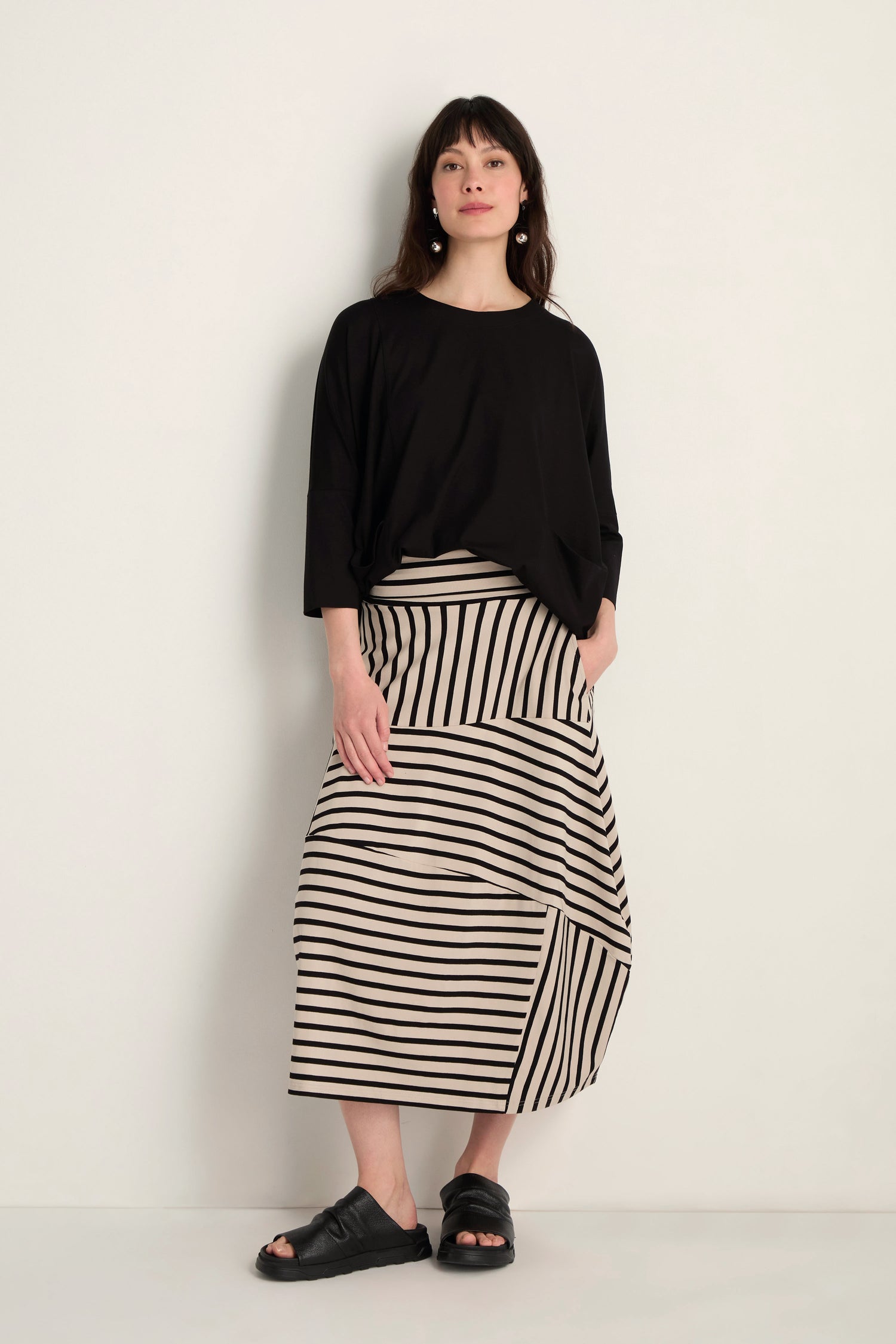 A woman stands against a plain wall wearing a loose black top, black sandals, and the Stripe Jersey Bubble Skirt—a midi-length skirt with stripes and a bubble silhouette.