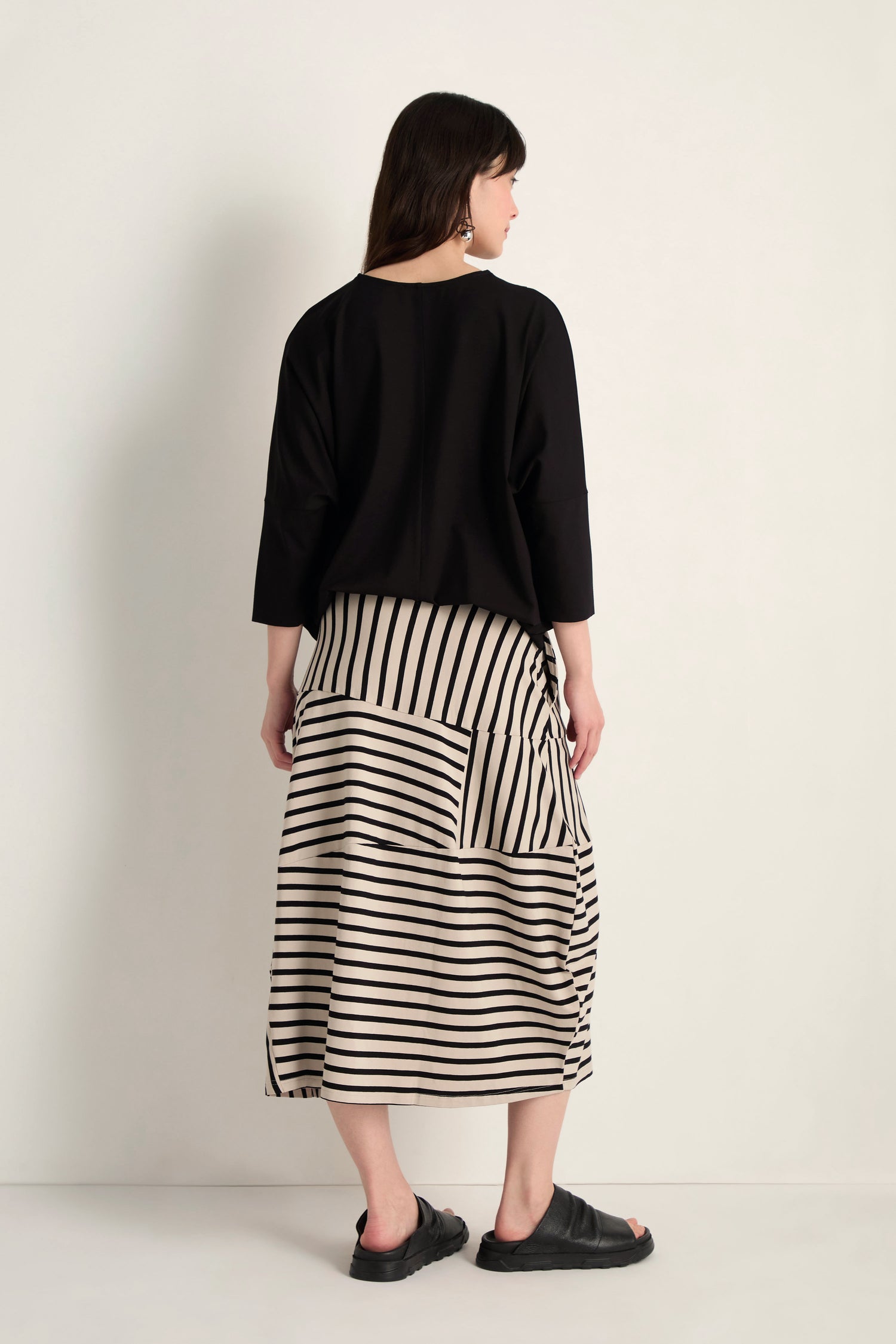 A woman faces away, wearing a black top with three-quarter sleeves, the Stripe Jersey Bubble Skirt in a midi striped bubble silhouette, and black sandals.