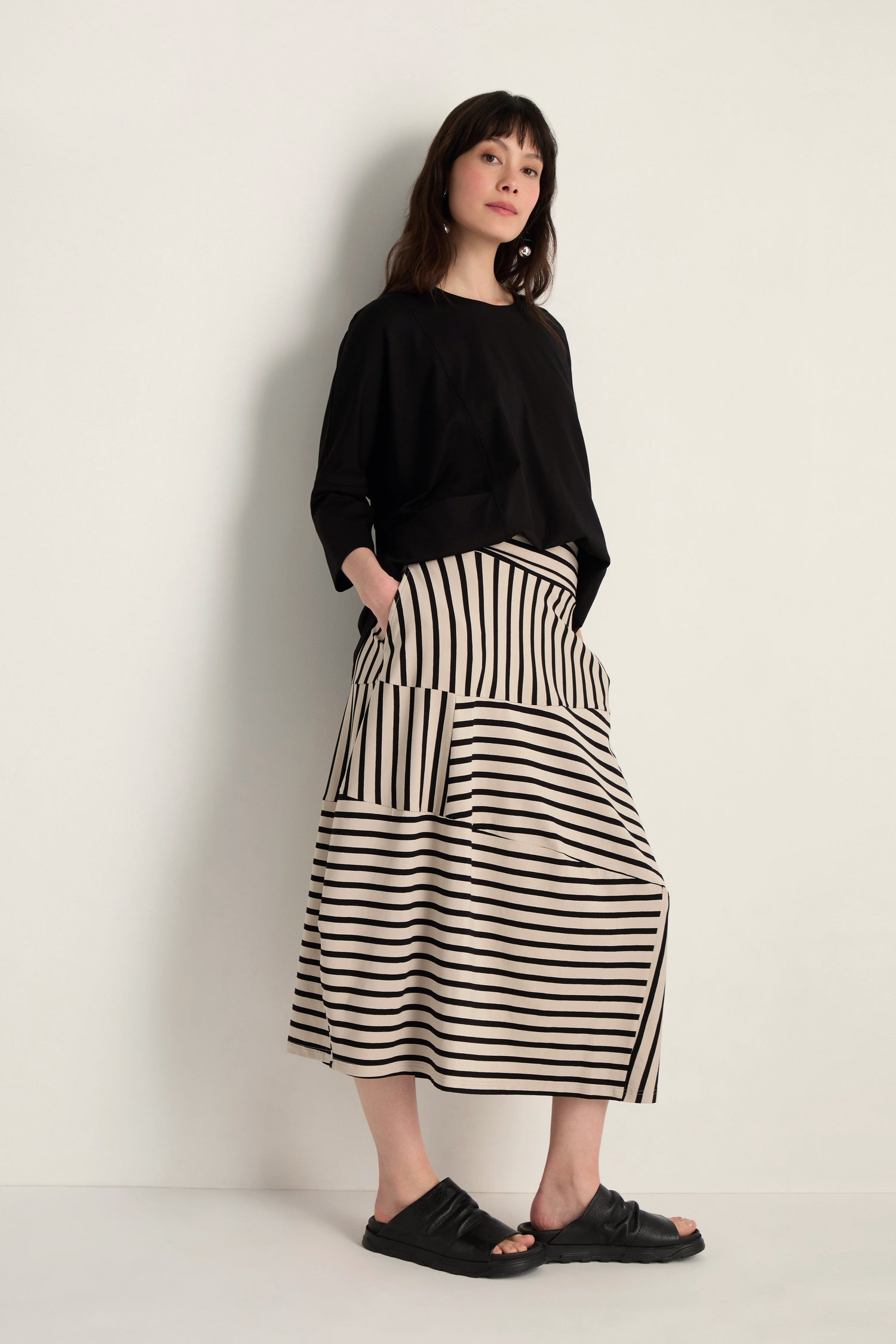 Woman stands against a light wall in a loose black top, Stripe Jersey Bubble Skirt with cream and black stripes plus pockets, and black slide sandals.