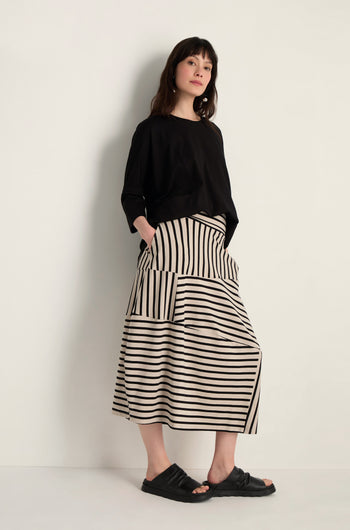 Woman stands against a light wall in a loose black top, Stripe Jersey Bubble Skirt with cream and black stripes plus pockets, and black slide sandals.