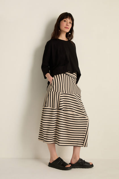 Woman stands against a light wall in a loose black top, Stripe Jersey Bubble Skirt with cream and black stripes plus pockets, and black slide sandals.