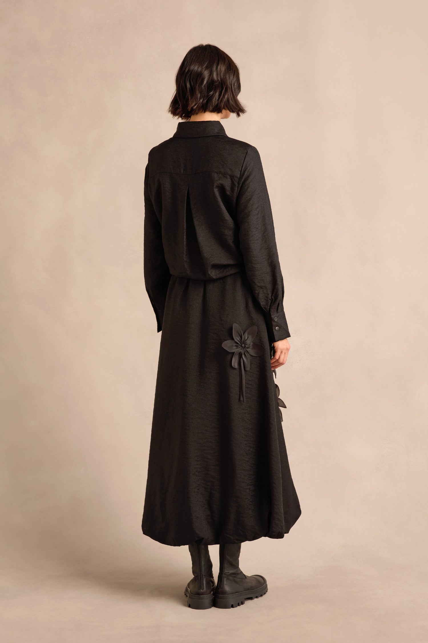 A person stands with their back to the camera against a beige background, wearing the Flower Detail Bubble Skirt—a long black midi-length skirt featuring floral appliqué details—paired with black boots.
