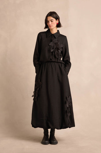 A woman stands against a plain neutral background with her hands in the pockets of a long-sleeved black Flower Detail Bubble Skirt midi dress, featuring large fabric floral appliqués.