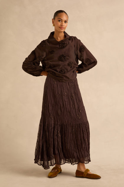 A person wears a dark brown long-sleeved top with floral appliqués and a matching crinkled chocolate Sheer Lined Pleated Skirt, paired with brown flats.