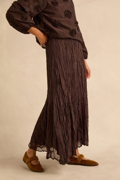 A person wearing a dark brown crinkled Sheer Lined Pleated Skirt and a matching long-sleeved top, paired with brown velvet loafers.