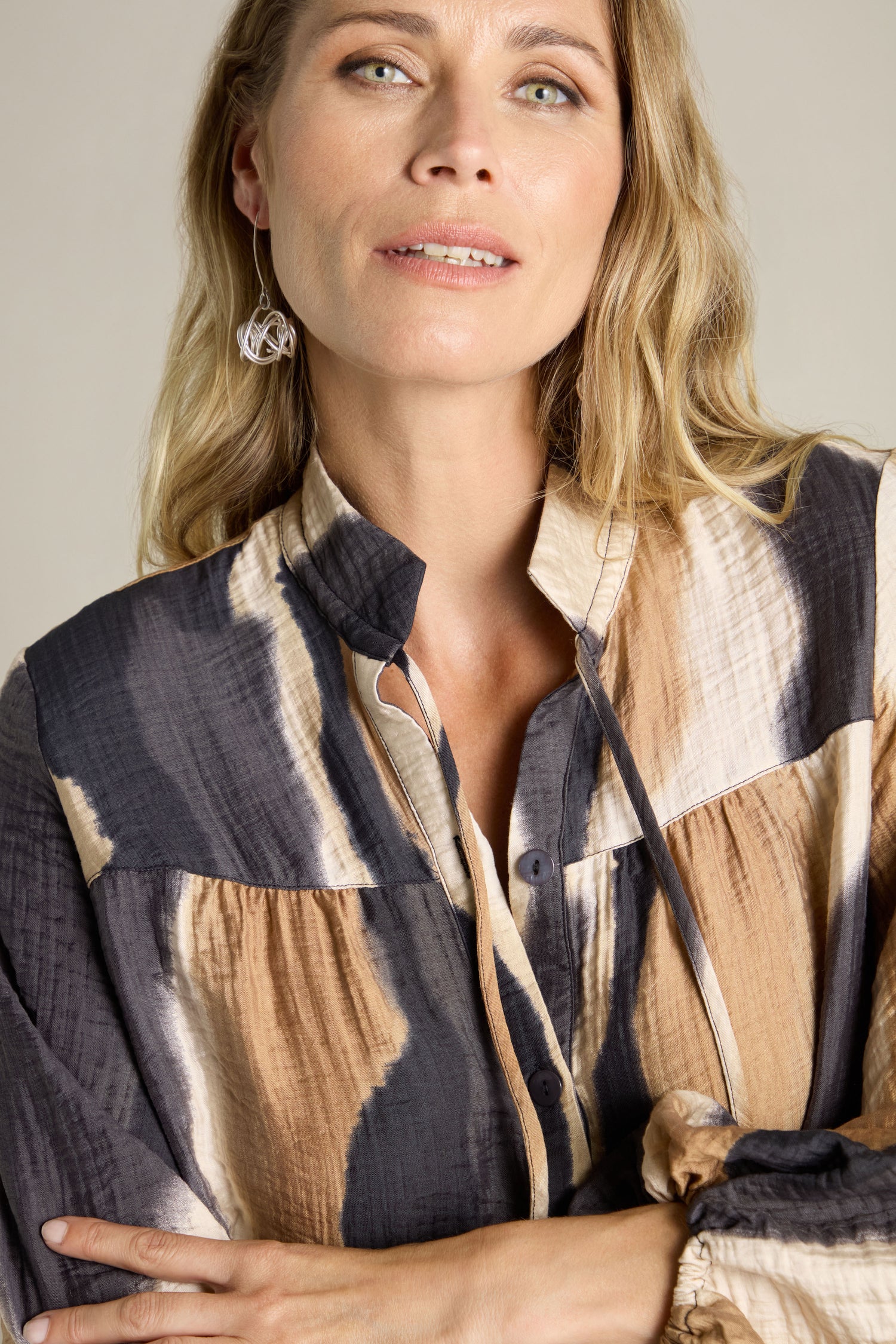 A person with blonde hair, wearing a black and beige Crinkle Viscose Print Shirt and geometric earrings, looks at the camera against a plain backdrop.