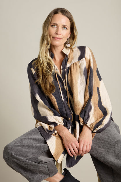 A woman with long, light brown hair, wearing the Crinkle Viscose Print Shirt in a beige and black pattern with a tie collar and gray pants, is seated on a stool against a plain background.