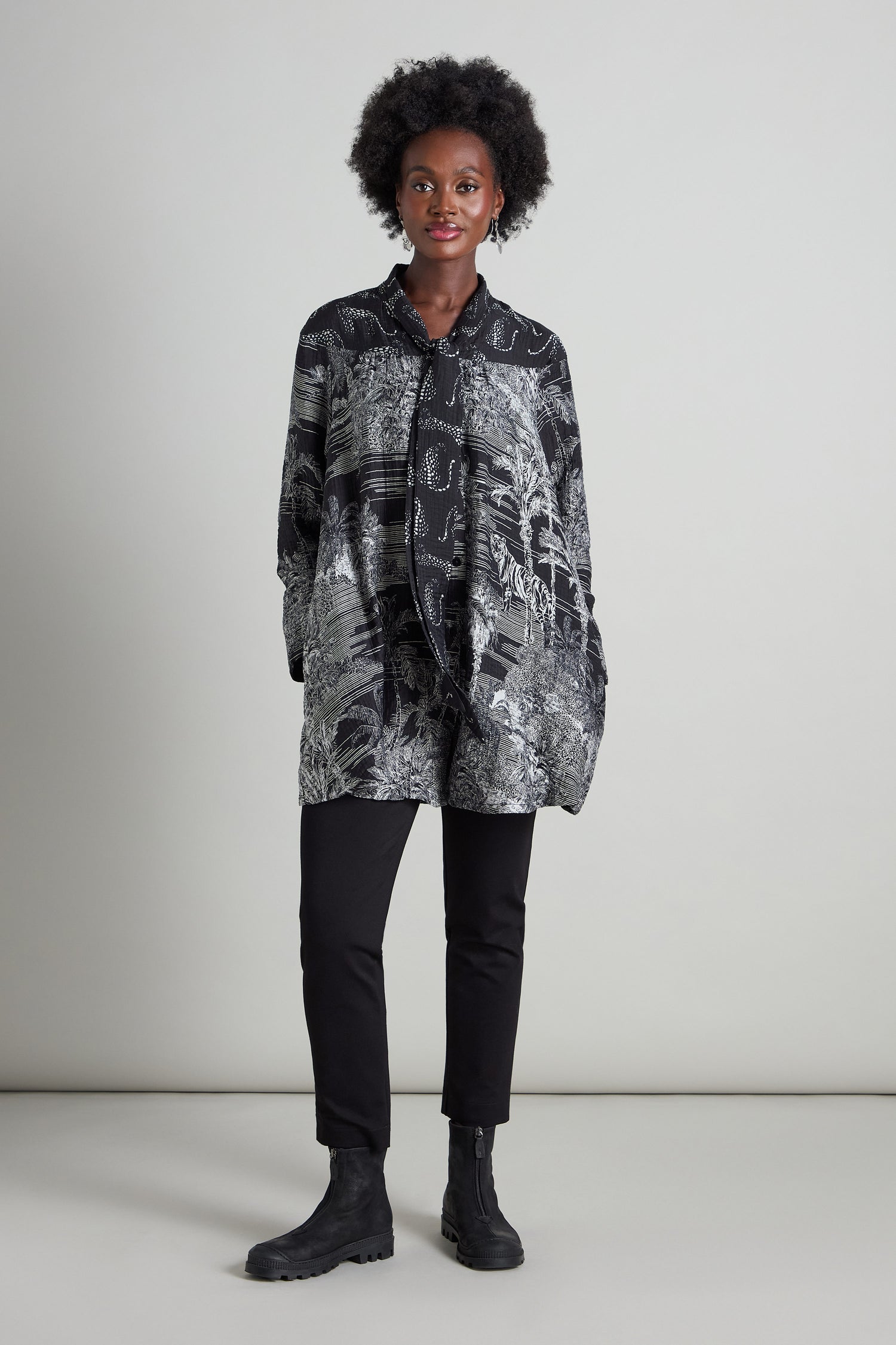 A person stands against a plain background, wearing a jungle print Tie Neck Viscose Shirt from Noen, paired with black pants and black boots.