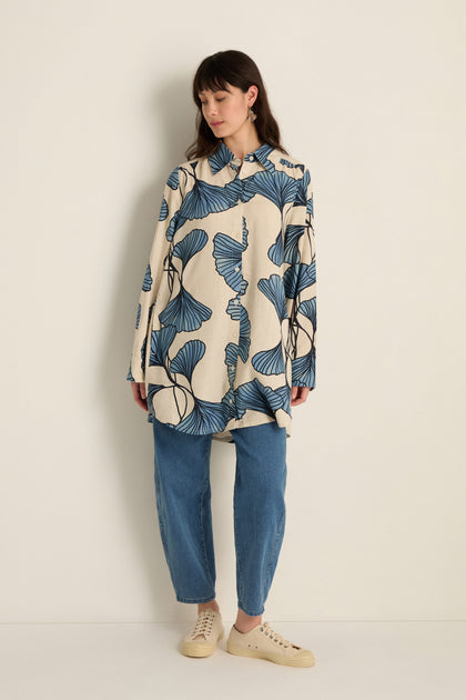 A woman stands against a plain wall, wearing the Ginkgo Viscose Linen Shirt with a blue ginkgo print, paired with blue jeans and beige sneakers.