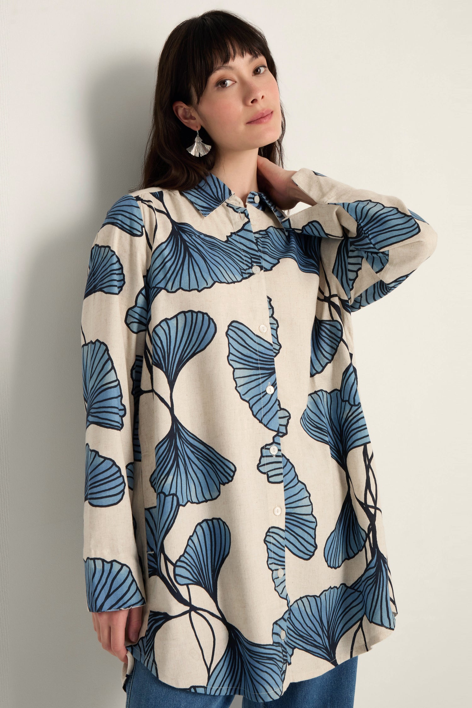 A woman stands against a plain wall wearing the Ginkgo Viscose Linen Shirt, which features a bold blue ginkgo leaf print on a light background and offers a relaxed silhouette.