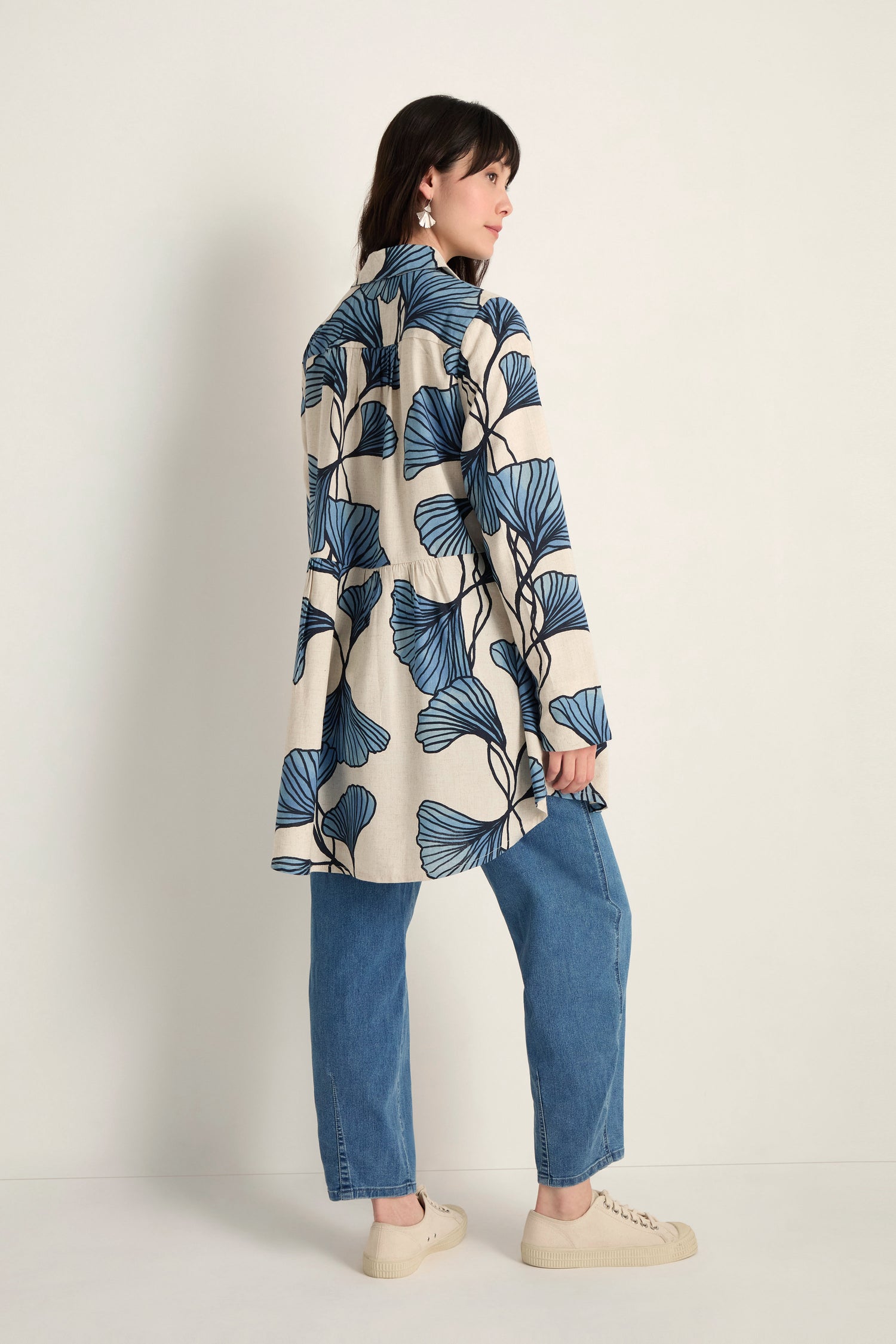 A person faces away, wearing the Ginkgo Viscose Linen Shirt—long, cream-colored with a blue leaf print—paired with blue jeans and beige shoes against a plain background.