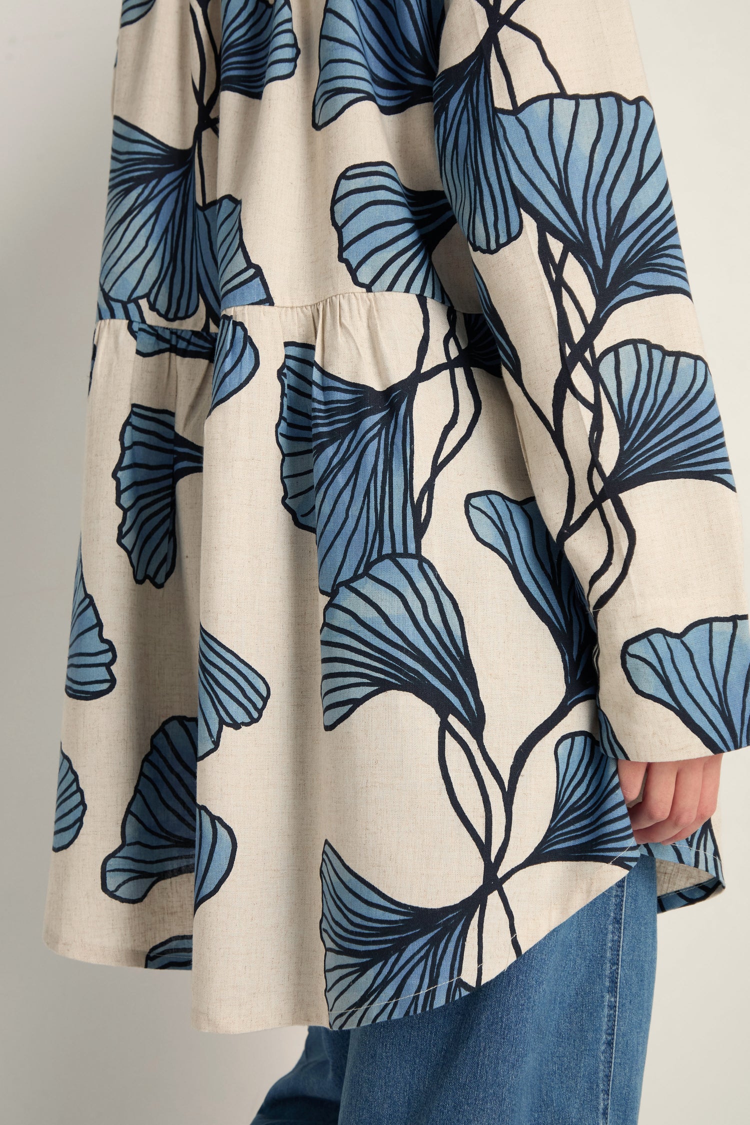 A person in the Ginkgo Viscose Linen Shirt—beige with large blue leaf designs—in a relaxed silhouette, paired with blue jeans, shown from shoulders to thighs.