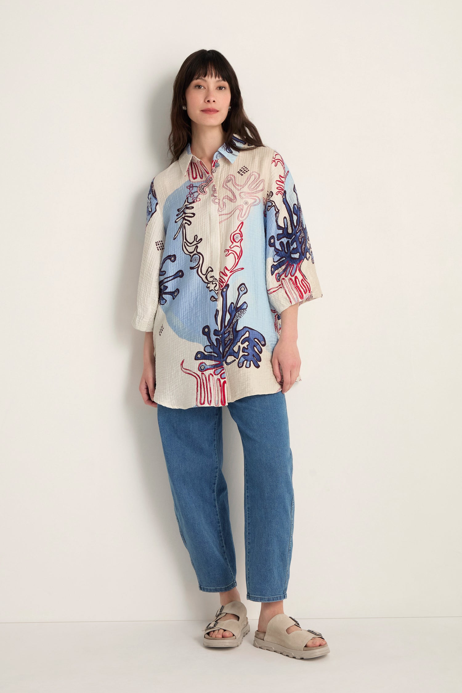 A woman stands against a plain wall wearing the Abstract Botanical Crinkle Shirt with blue and red designs, paired with blue jeans and beige sandals.