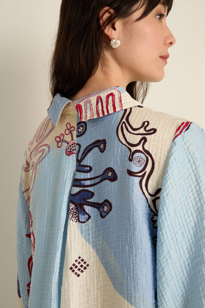 A woman with long dark hair and silver earrings is shown from the side and back wearing the Abstract Botanical Crinkle Shirt, featuring light blue and cream hues with Scandinavian-inspired abstract botanical designs.
