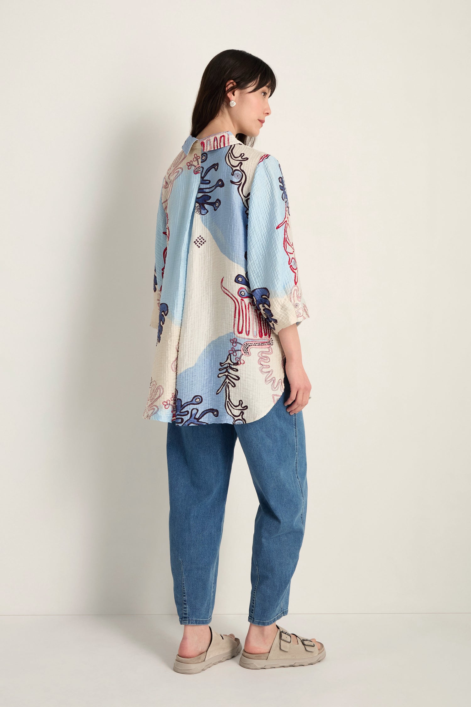 A woman stands sideways against a plain light background, wearing the Abstract Botanical Crinkle Shirt with blue, cream, and red designs, paired with blue jeans and beige shoes.