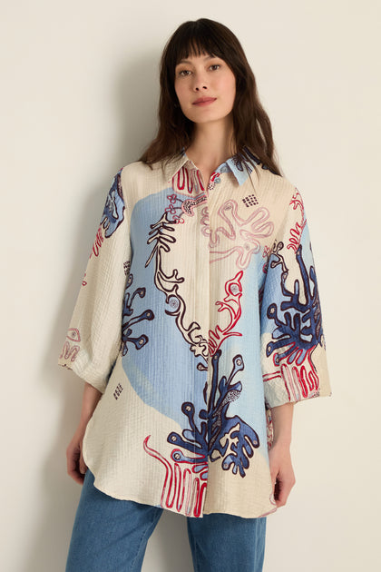 A woman stands against a plain wall, wearing the Abstract Botanical Crinkle Shirt featuring loose, long sleeves and red, blue, and beige Scandinavian-style patterns, casually paired with blue jeans.