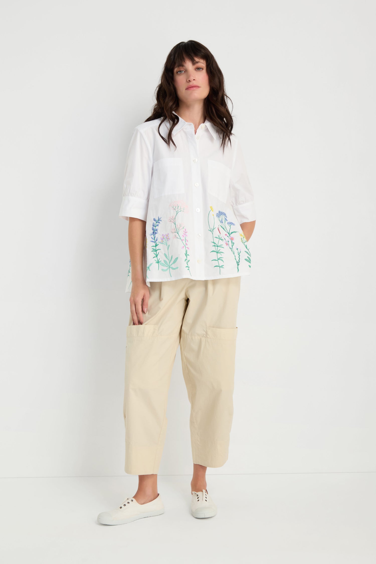 A woman stands against a white background wearing the Embroidered Hem Shirt, featuring crisp cotton with floral embroidery, paired with beige wide-leg pants and white sneakers.