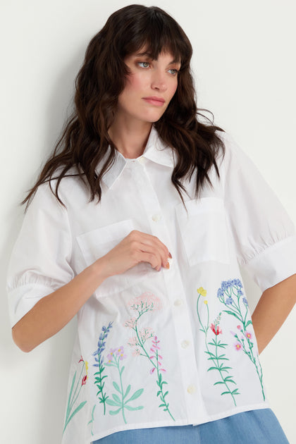 A woman in the Embroidered Hem Shirt, a crisp cotton short-sleeve button-up with multicolored floral embroidery, stands against a white background.