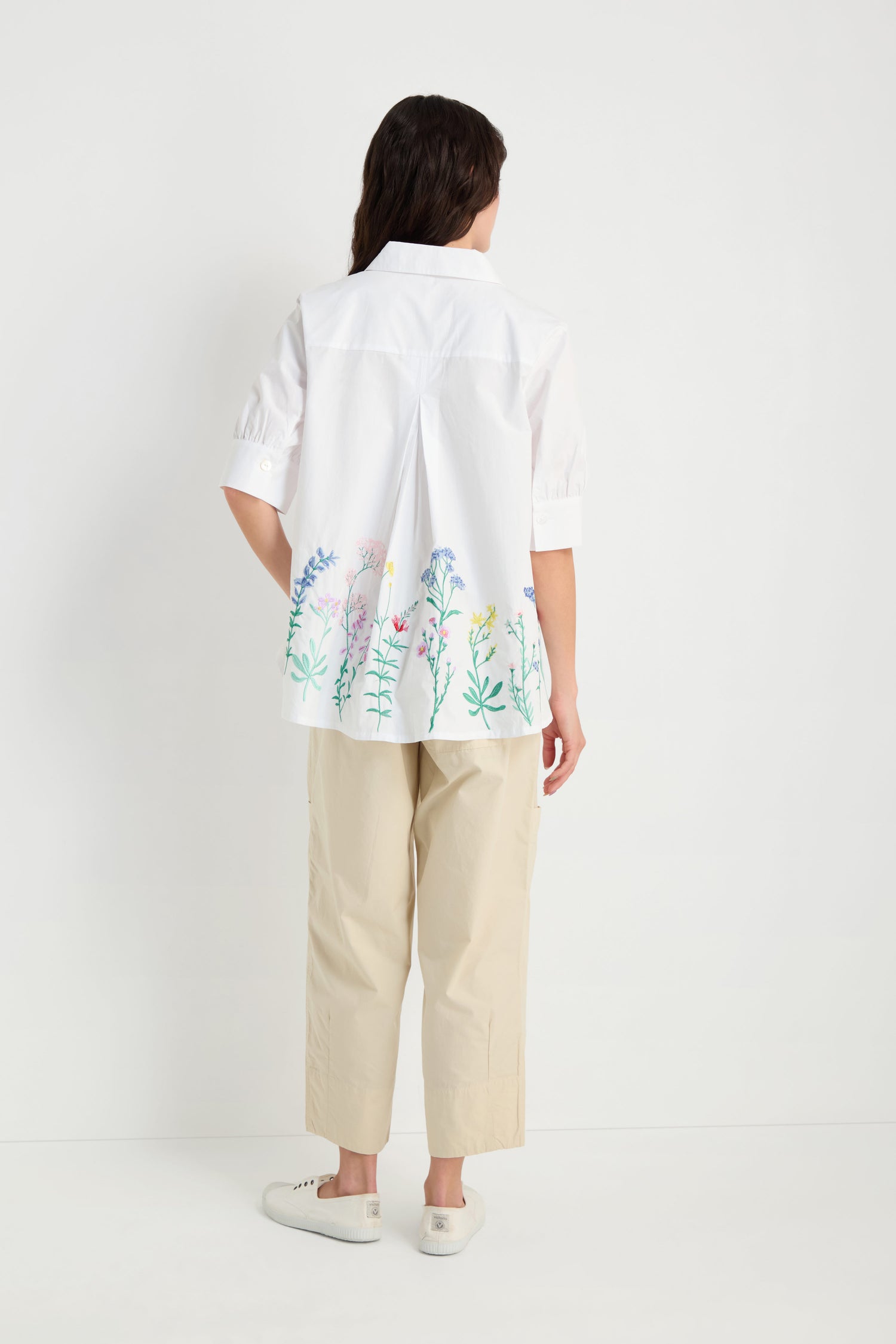 A person stands facing away, wearing the Embroidered Hem Shirt—a crisp cotton shirt with floral details—paired with beige pants and white sneakers against a plain white background.