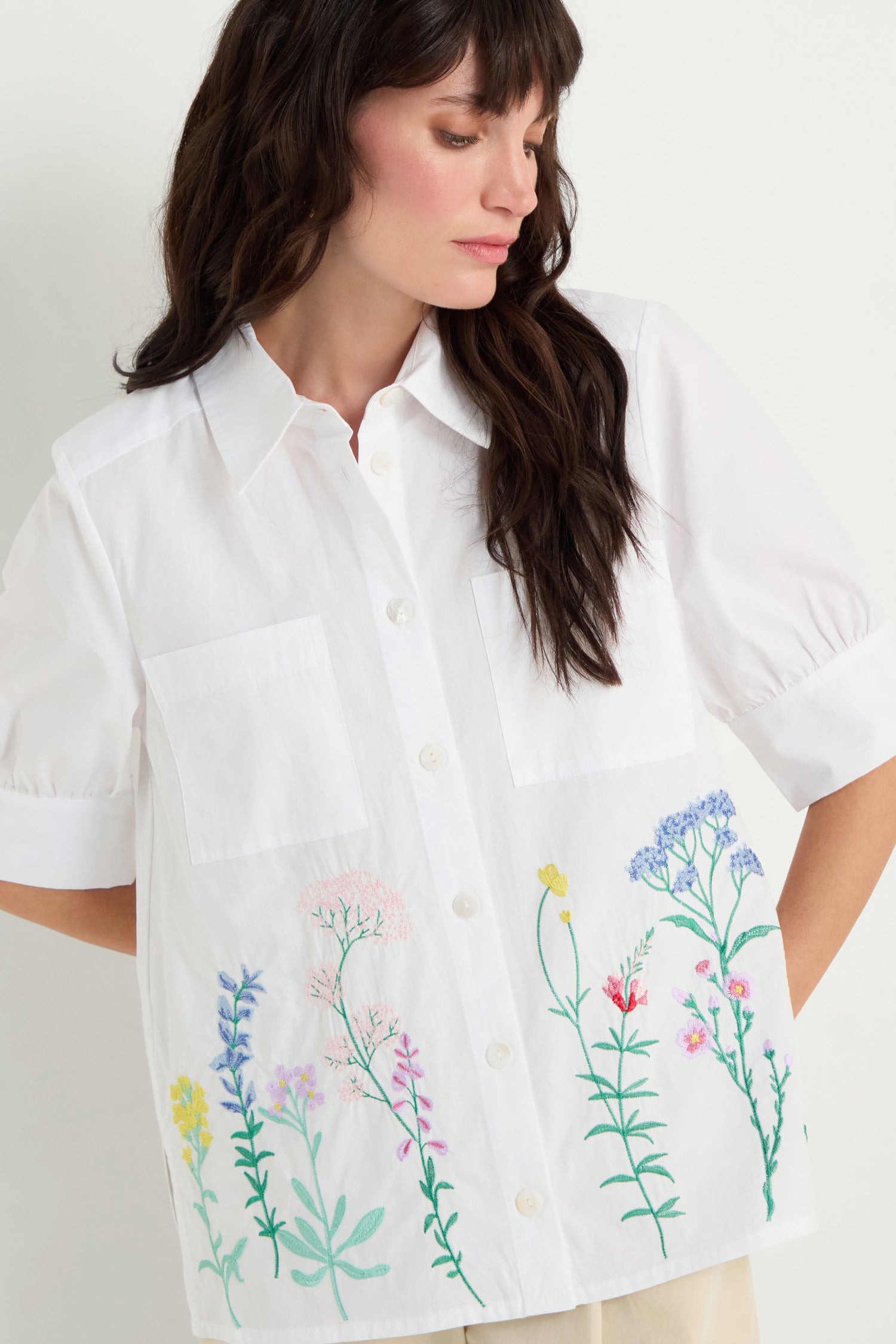 A woman wears the Embroidered Hem Shirt—a crisp cotton, short-sleeve button-up featuring two chest pockets and colorful floral embroidery along the hem.
