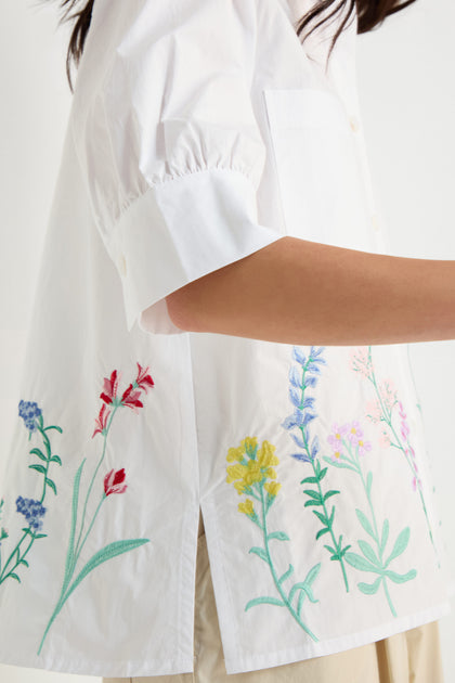 A person wearing the Embroidered Hem Shirt, featuring crisp cotton fabric and multicolored floral embroidery near the hem, shown from shoulder to hip.