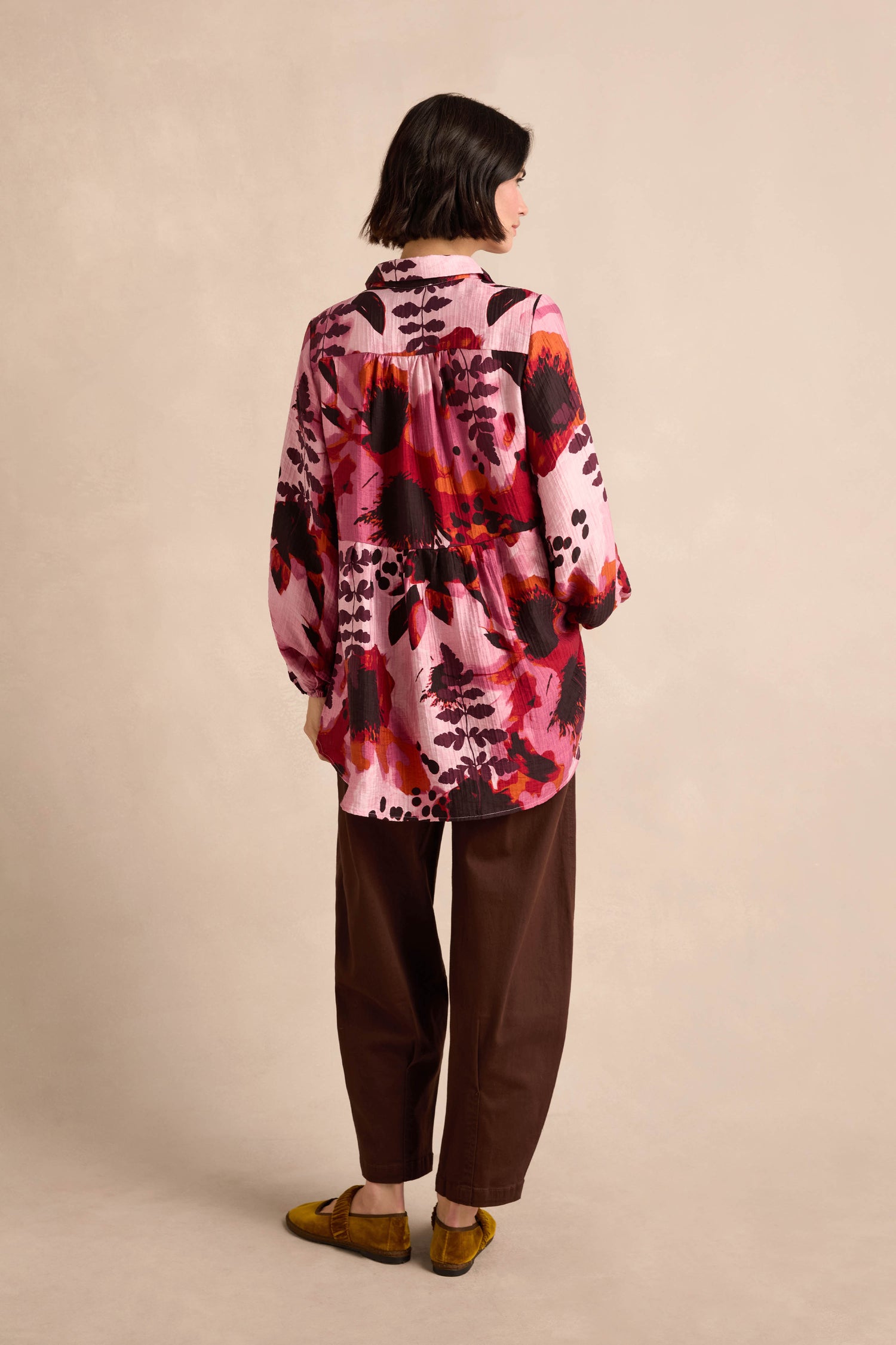 A person stands with their back to the camera, wearing the Crinkle Viscose Shirt in a pink and red botanical print, paired with brown pants and yellow shoes against a neutral background.
