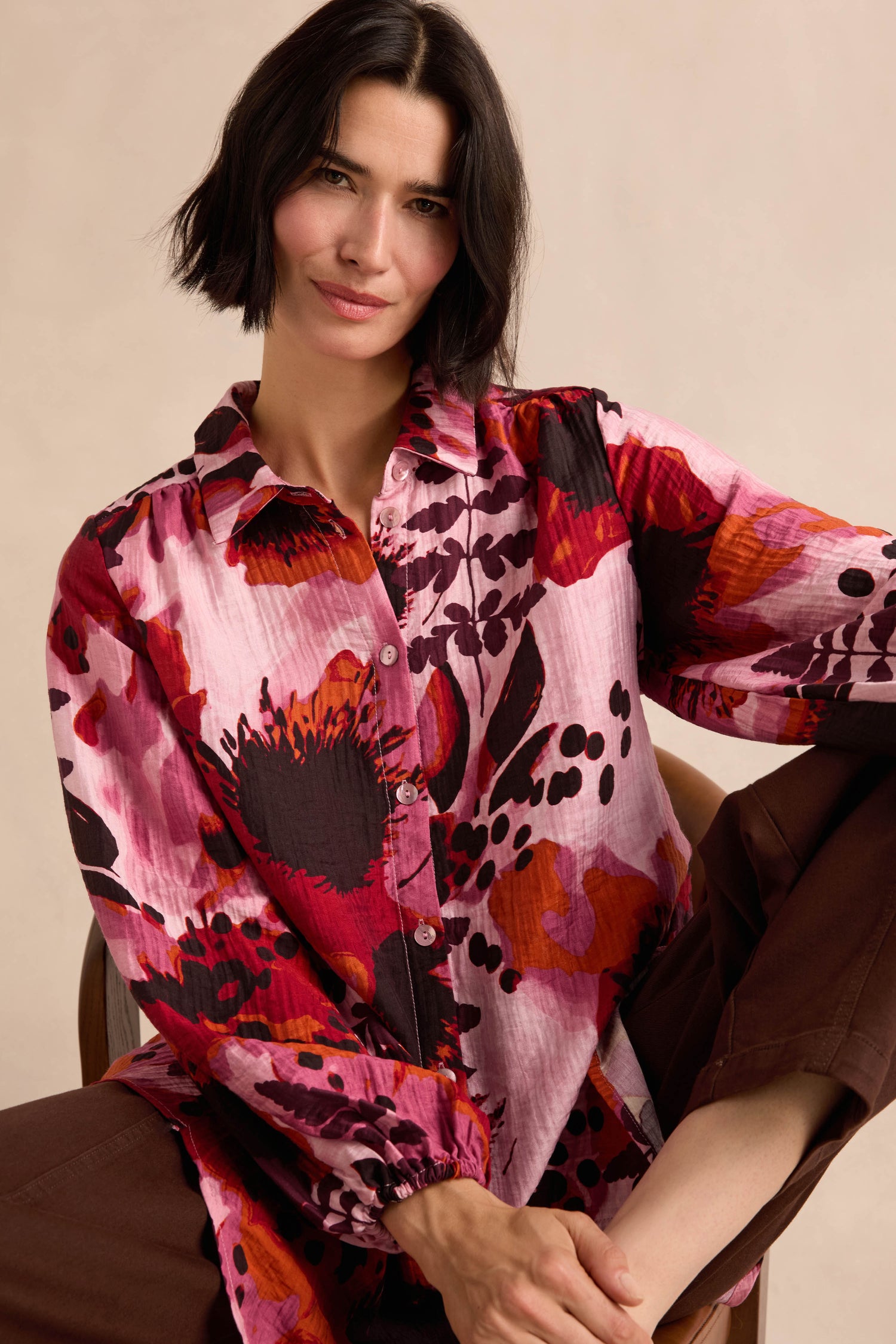 A woman with a bob haircut is seated with one arm resting on her knee, wearing the Crinkle Viscose Shirt—a long-sleeve, button-up top featuring a bold pink and red botanical print.